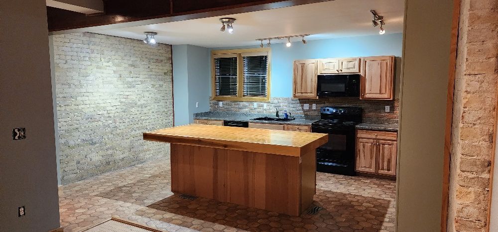 Kitchen with wood cabinets, black appliances, and a butcher block island. Brick and wood flooring.