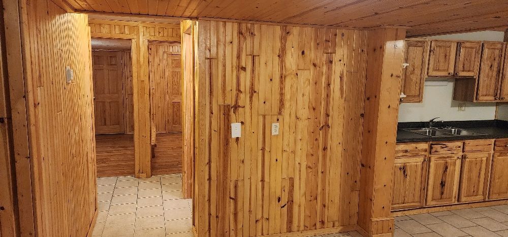 Interior view of a kitchen and doorway. Walls are wood paneling. Cabinets and floor are also wood.