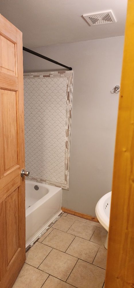 Bathroom with a bathtub, shower curtain, toilet, and tan tile floor. Light gray walls with a wooden door open.