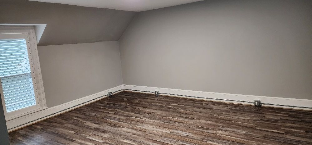 Empty room with gray walls and wood-look floor, slanted ceiling, window on the left.
