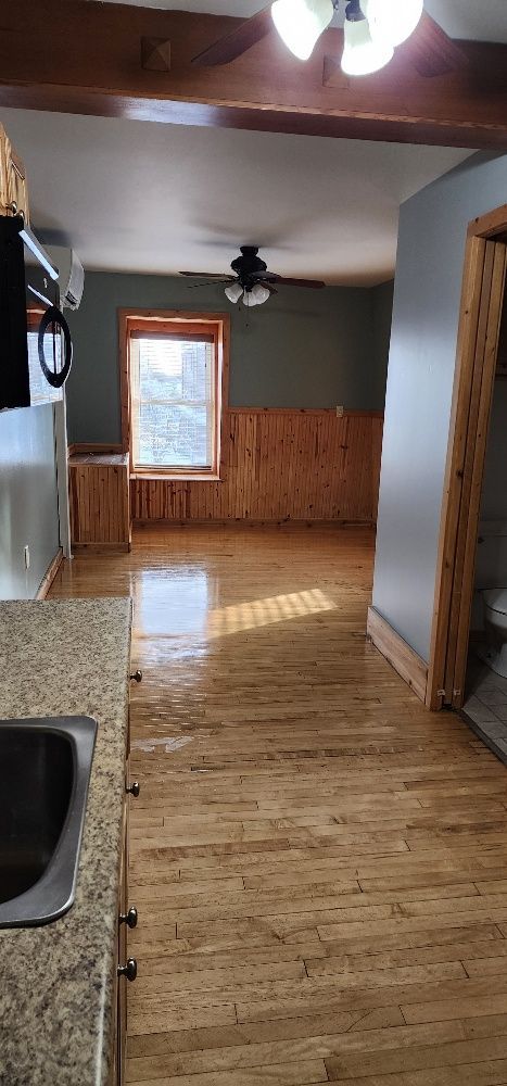 Interior view of a room with wood floors, walls, and ceiling beams; a window, ceiling fan, and kitchen counter are visible.