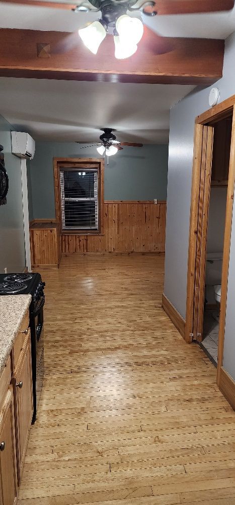 Interior view of a kitchen, showcasing wood floors, cabinets, and a doorway to a living space with a window.