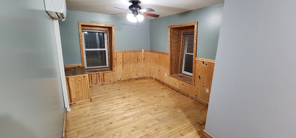 Room with wooden walls, light wood floor, and two windows. Blue walls, ceiling fan.