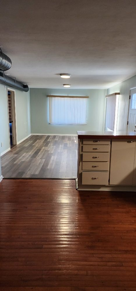 Interior shot of a room with wood flooring, kitchen cabinets, and a window with blinds.
