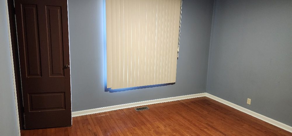 Empty room with hardwood floors, gray walls, brown door, and vertical blinds.
