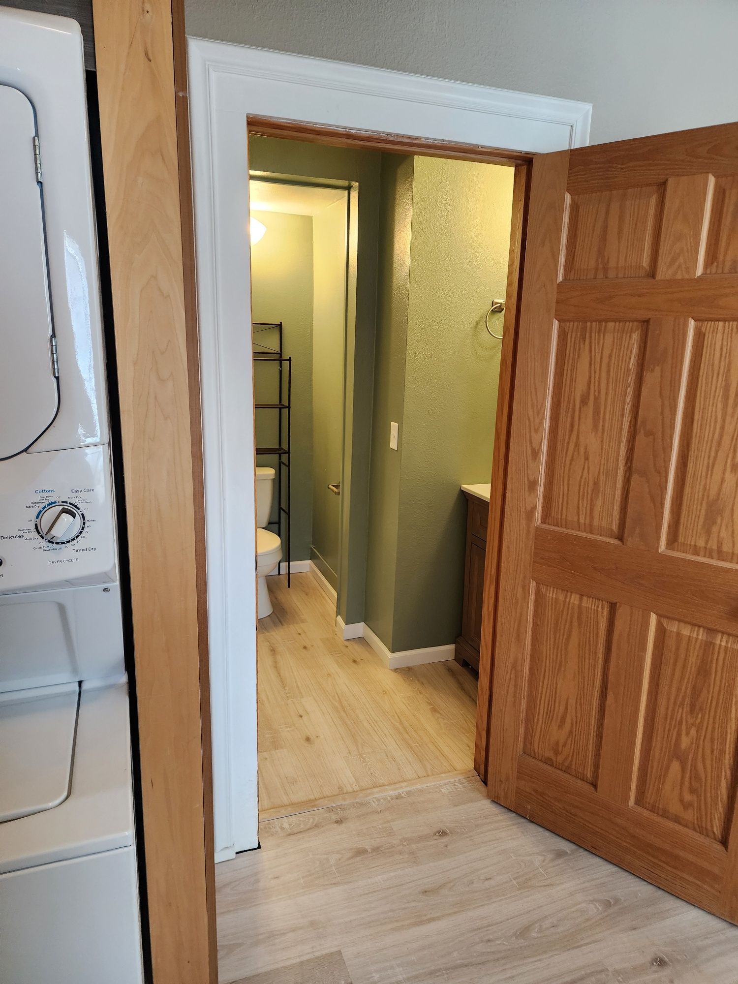 Laundry room and bathroom with wood-paneled door open to reveal toilet, sink, and stacked washer/dryer unit.