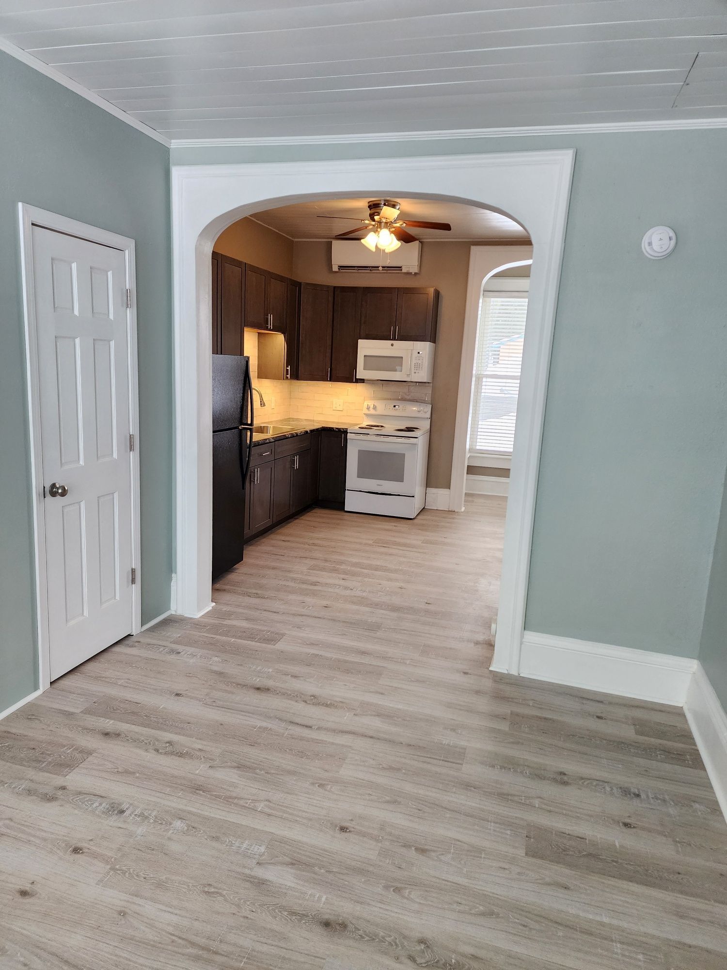 Interior view of a living space with a door on the left and a kitchen visible through an archway.