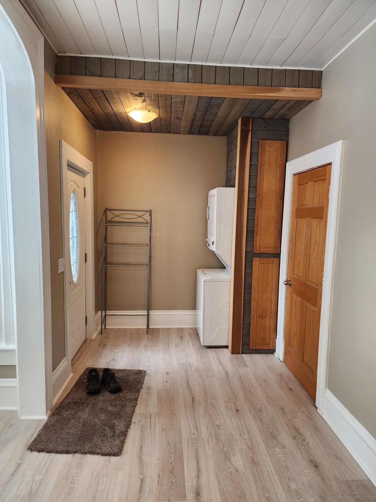 Entryway with washer/dryer, shelving, wooden door, and rug. Gray walls and flooring. White ceiling.