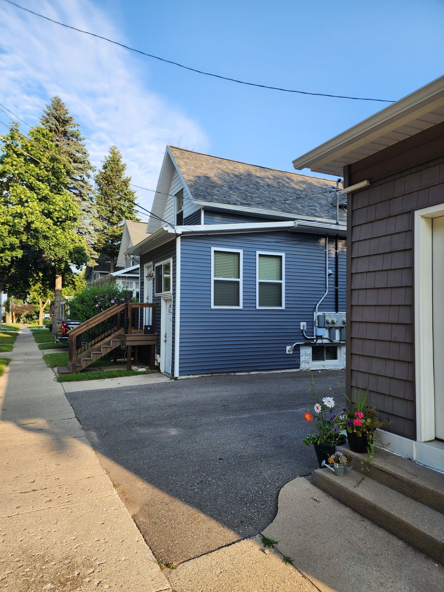 Sidewalk view of blue house, gray asphalt drive, and brown shingled structure. Flowers sit near entry stairs.