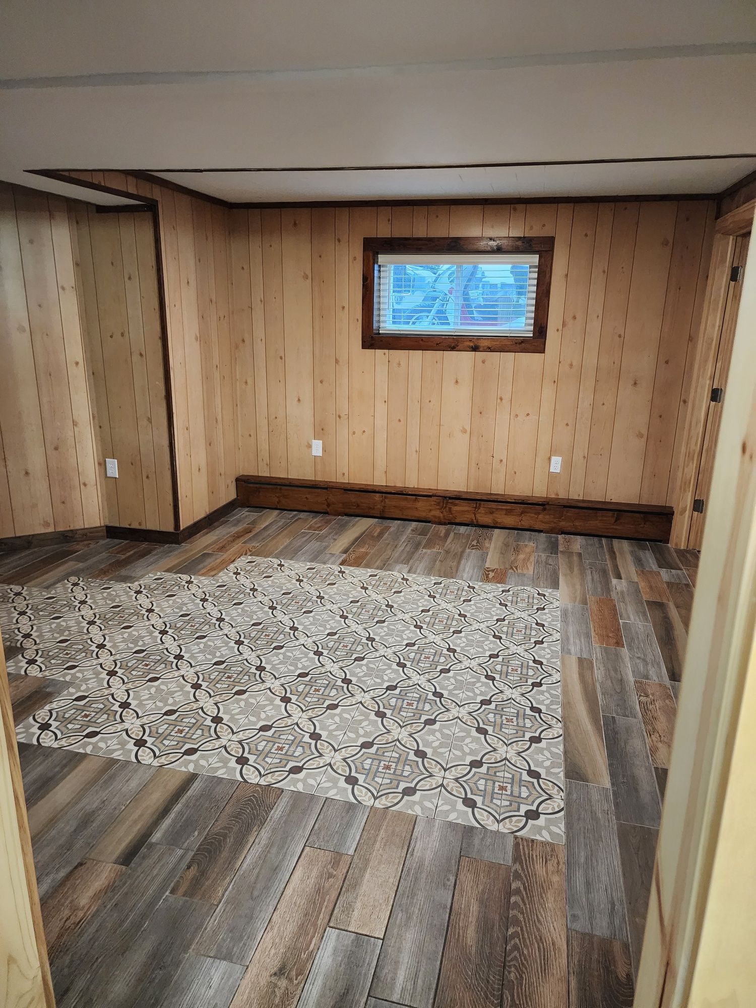 Room with wood paneling, wood-look flooring, area rug. Small window, brown trim.