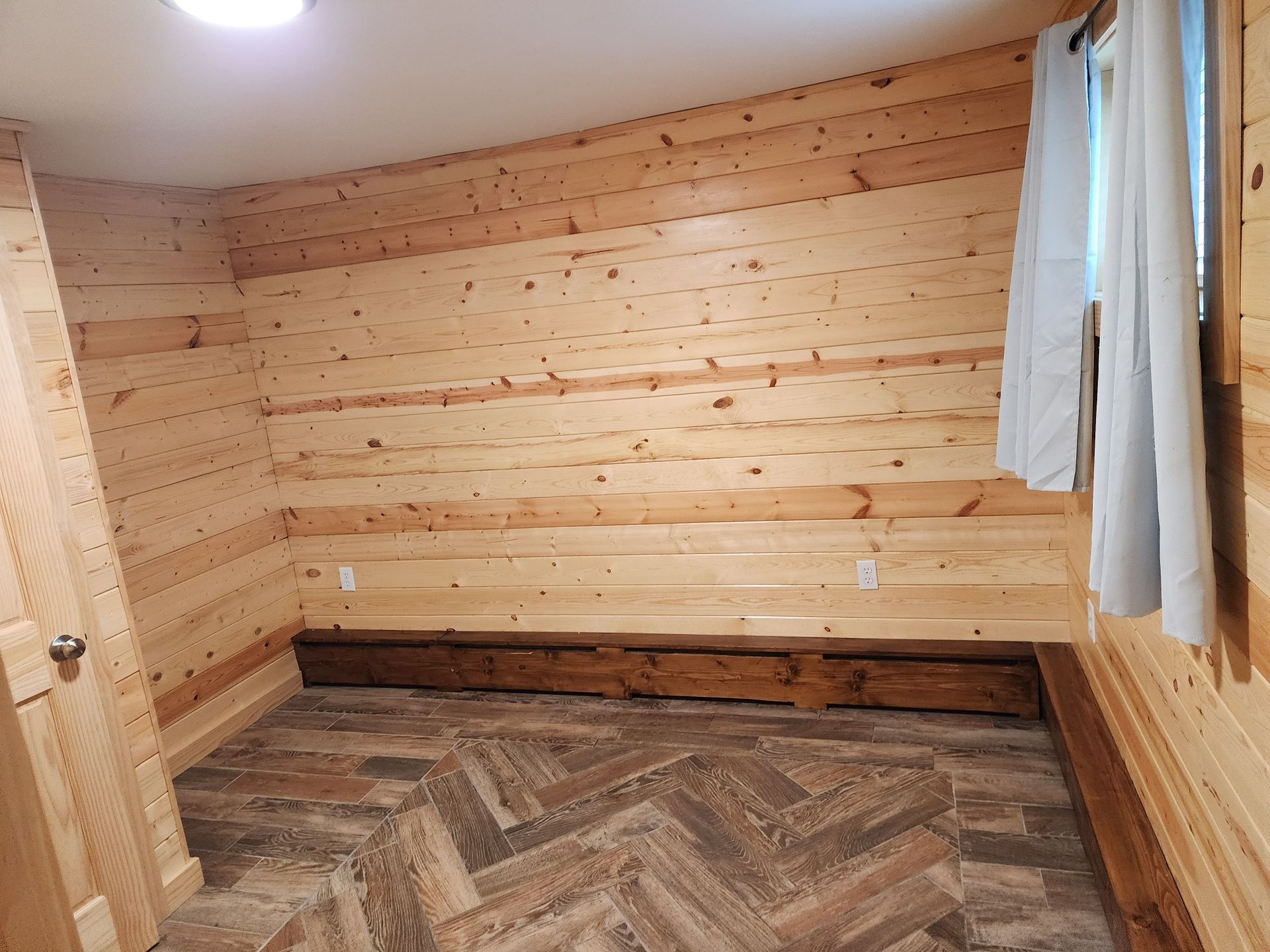 Empty room with wood paneled walls, a window with a curtain, and a herringbone floor.