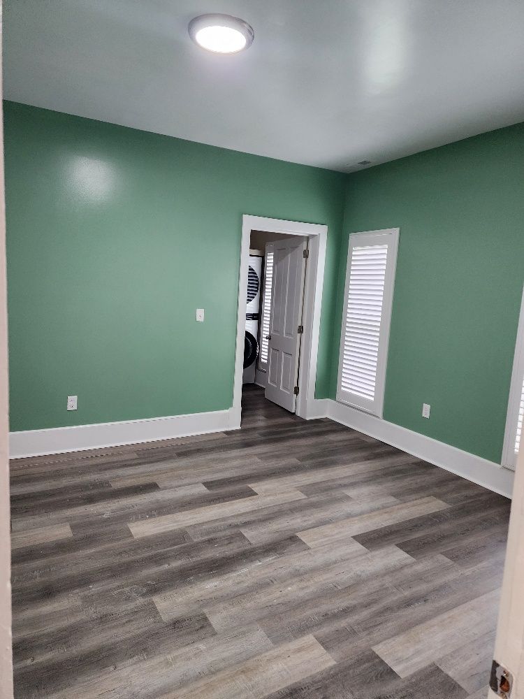Empty room with green walls, white trim, and gray wood-look flooring; doorway and window with shutters.