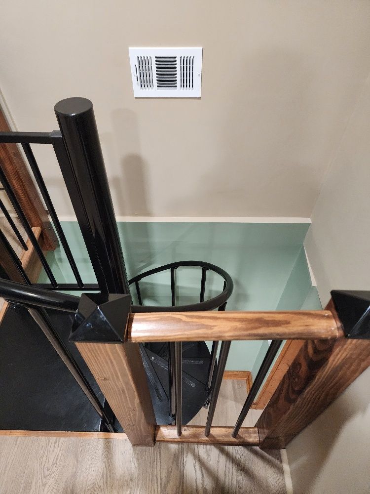 Spiral staircase with wood and black metal railings, leading downwards. Air vent on wall.