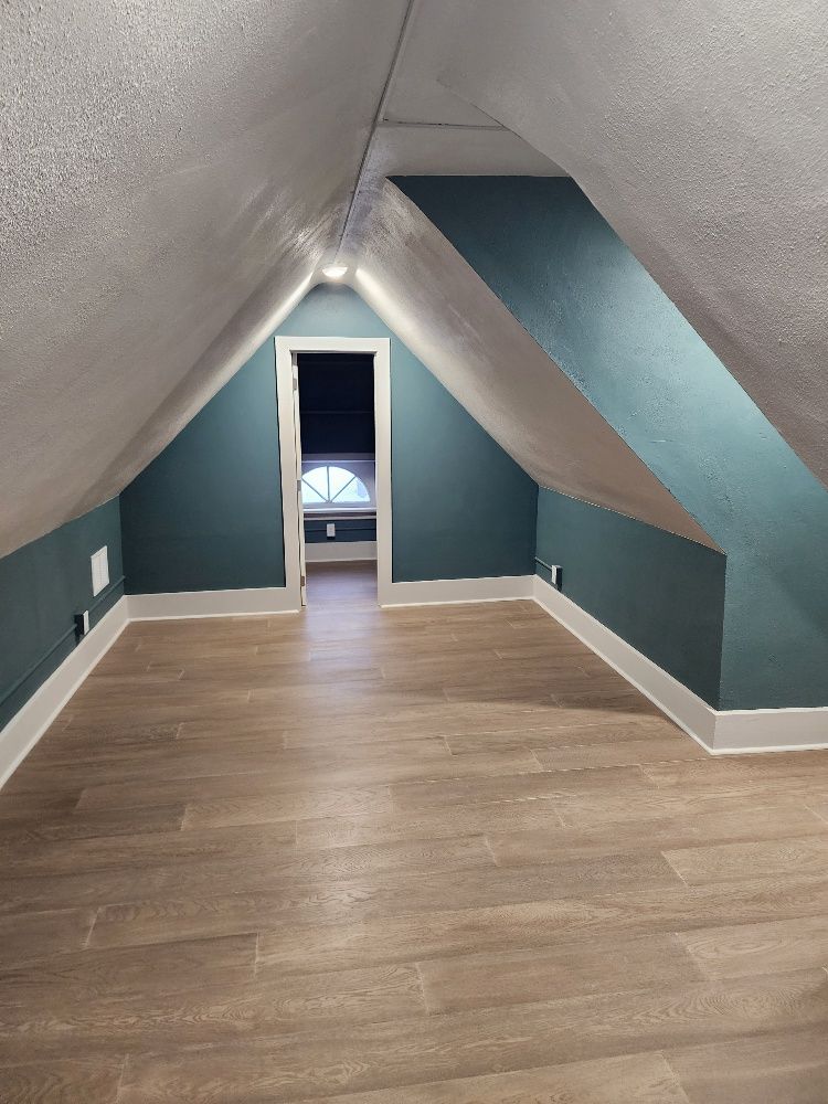 Attic room with sloped teal walls, white trim, and light wood-look flooring. Doorway leads to another space.
