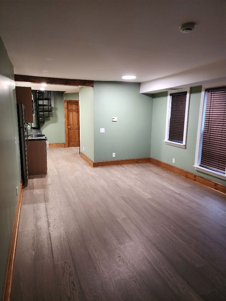 Empty apartment interior with wood flooring, green walls, and brown trim. Contains kitchen, spiral staircase, and windows with blinds.