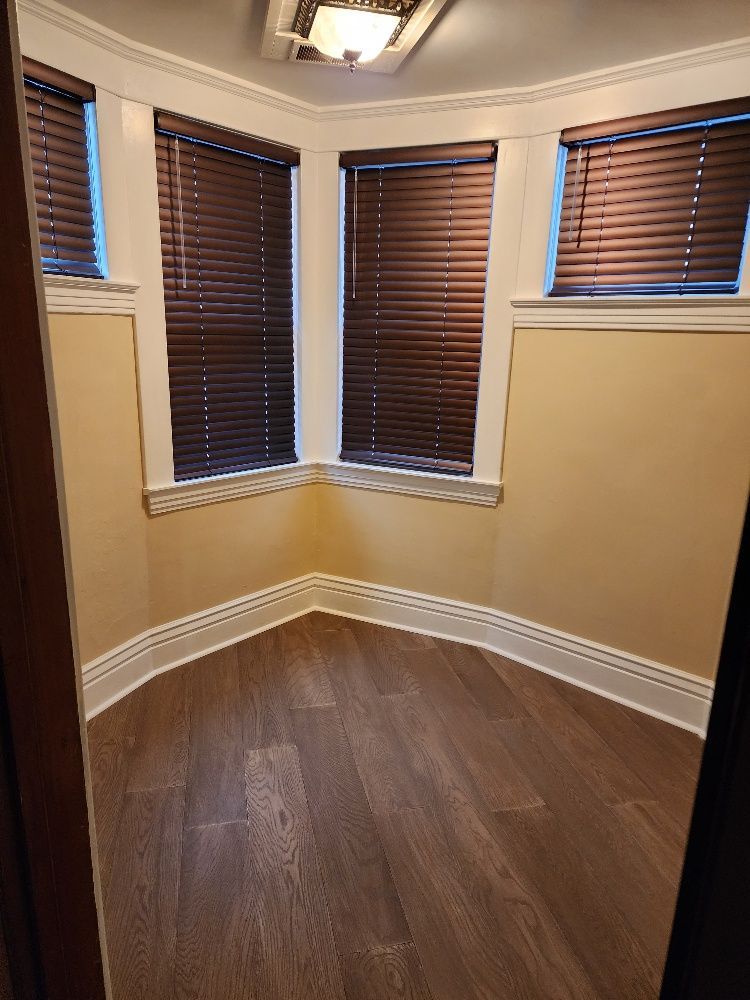 Empty room with dark wood floors, three windows with blinds, and beige walls.