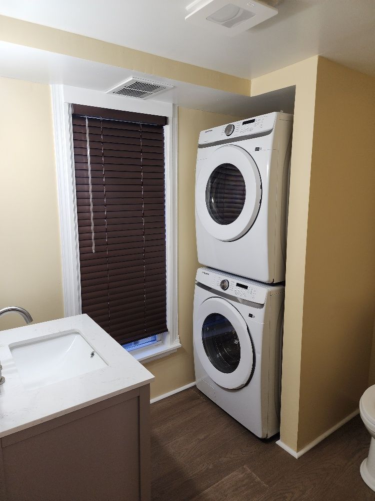 Stacked white washer and dryer in a small bathroom nook next to a window with brown blinds.
