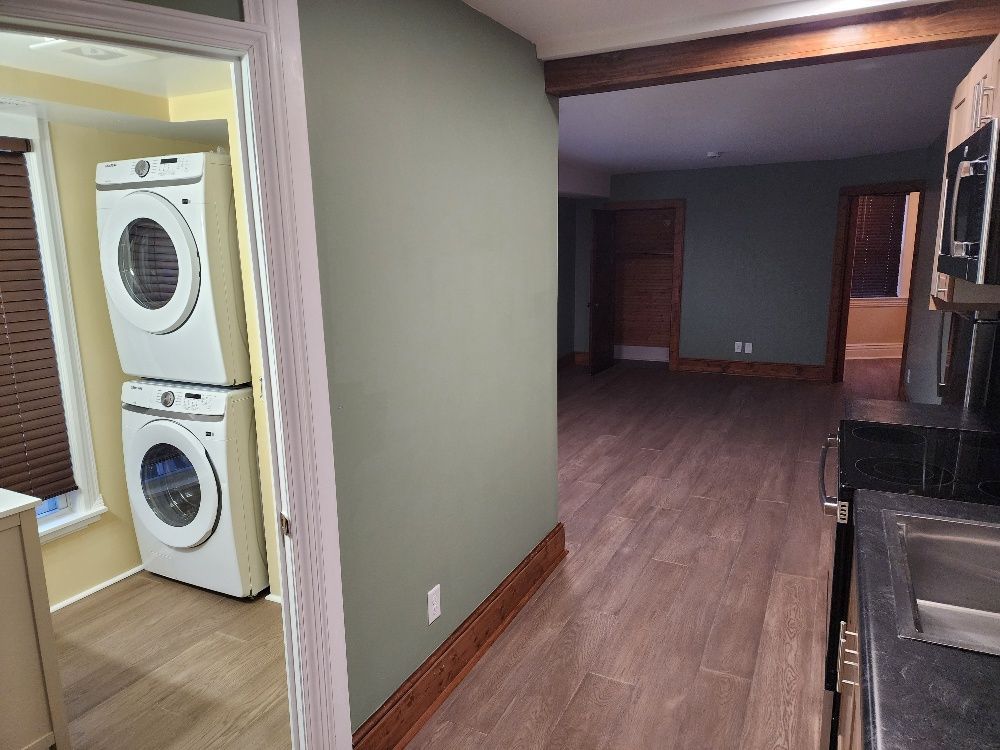 View into a house with laundry machines in a small room and an open space with wooden floors.