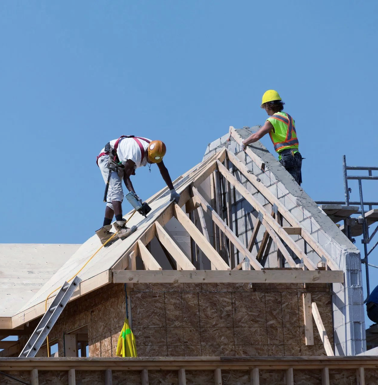 Deux ouvriers du bâtiment, casqués, travaillent sur la charpente d'un toit, construisant une maison sous un ciel bleu azur.
