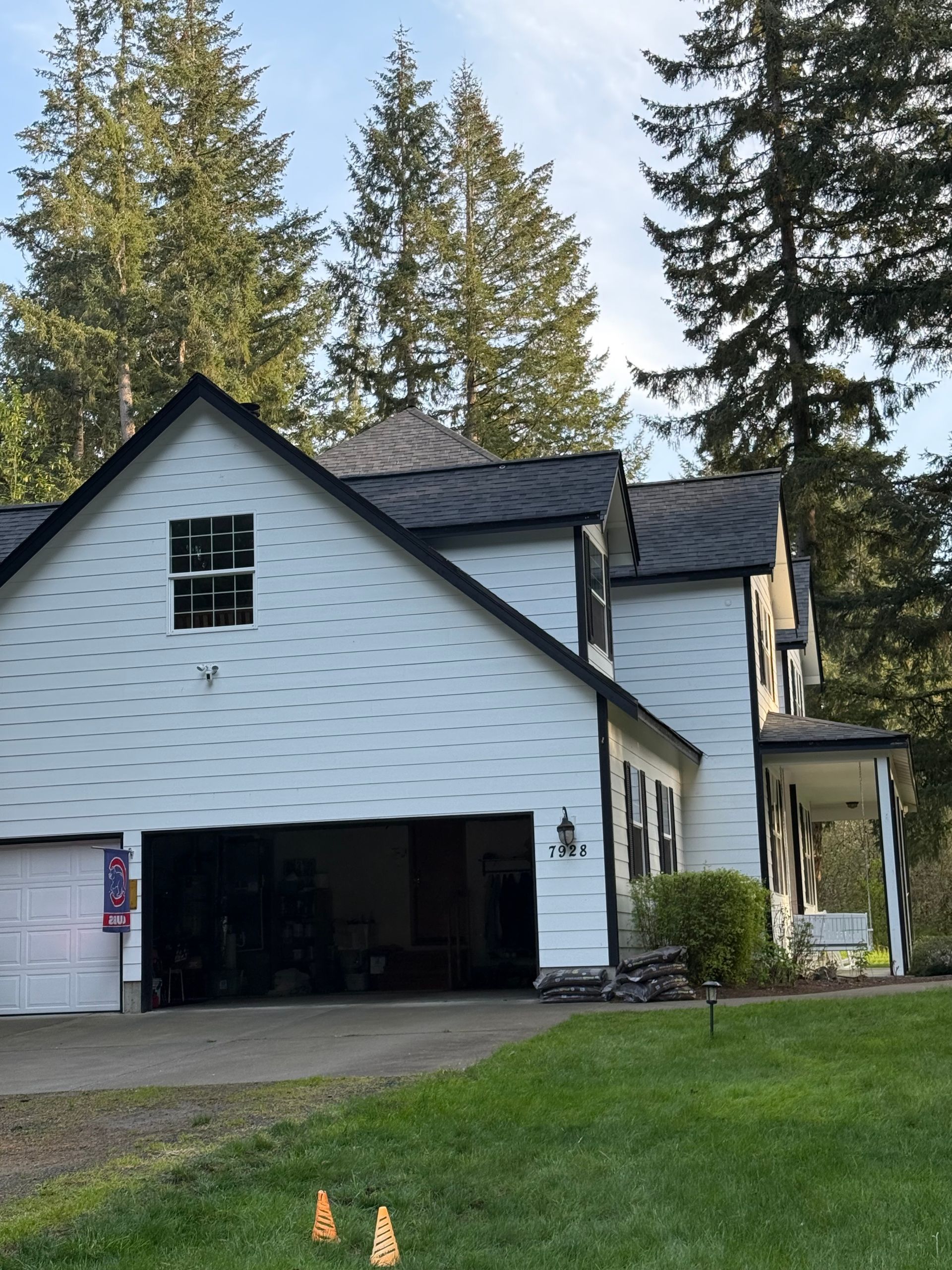 White house with black roof, open garage door, green lawn, and tall evergreen trees in the background.