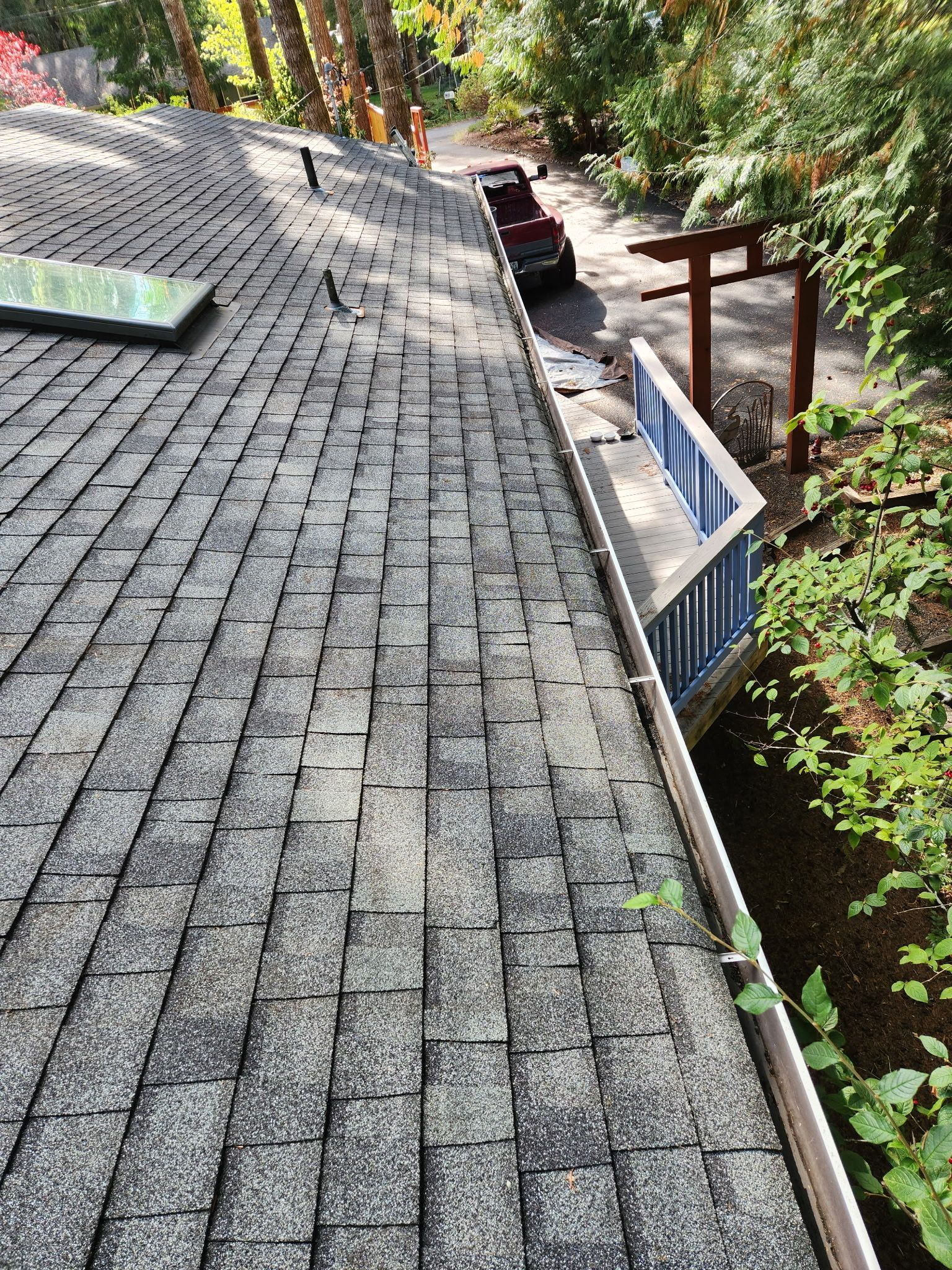 View of a roof with dark shingles, gutter, and a structure with blue and white accents, in a wooded setting.