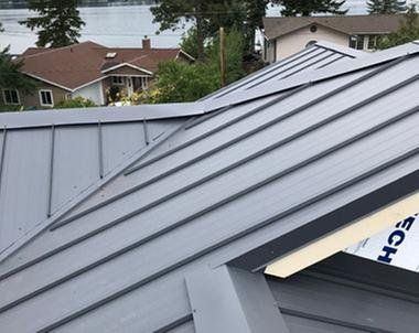 Gray metal roof with vertical seams, angled, with suburban homes in the background and a body of water in the distance.