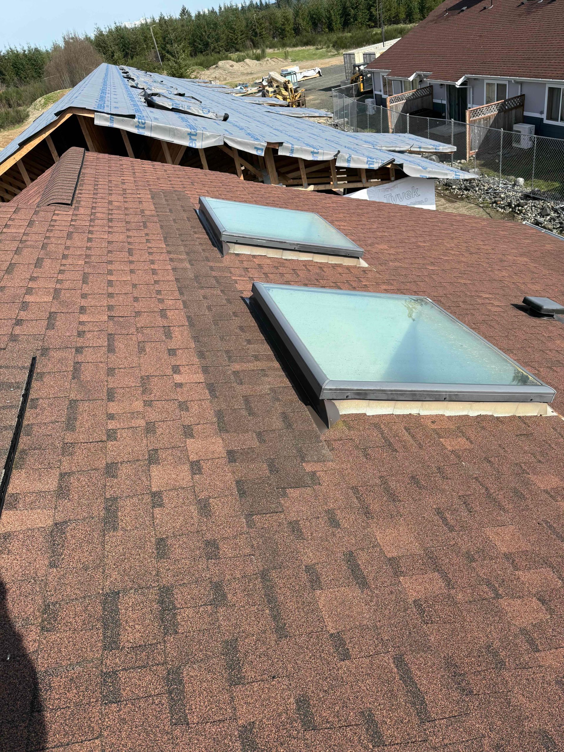 Roof with two skylights, red shingles, and damaged section in background.