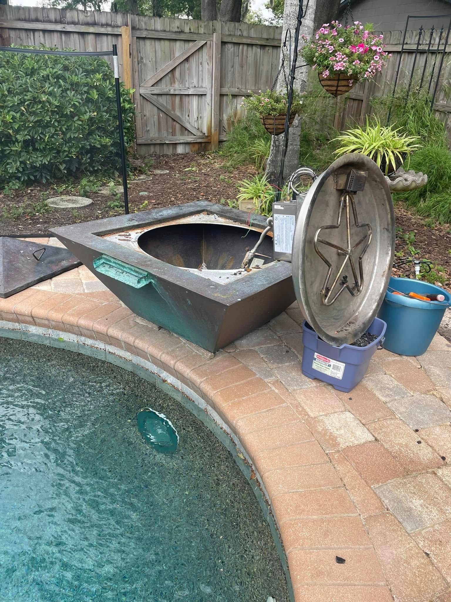 Outdoor fire pit next to a pool. Square, copper-colored pit with gas components exposed, on a brick patio.