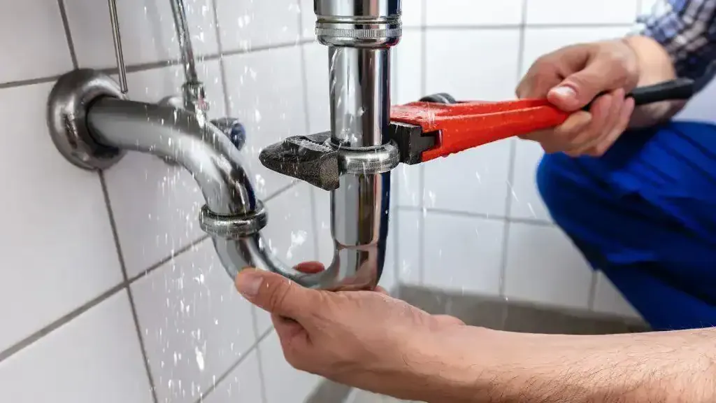 Plumber using a wrench to fix a leaking pipe in a bathroom, water spraying.