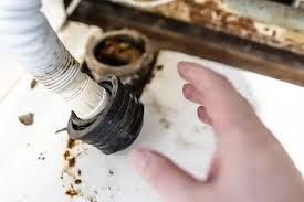 Hand reaching toward a clogged white pipe beneath a sink with dirt and debris around it