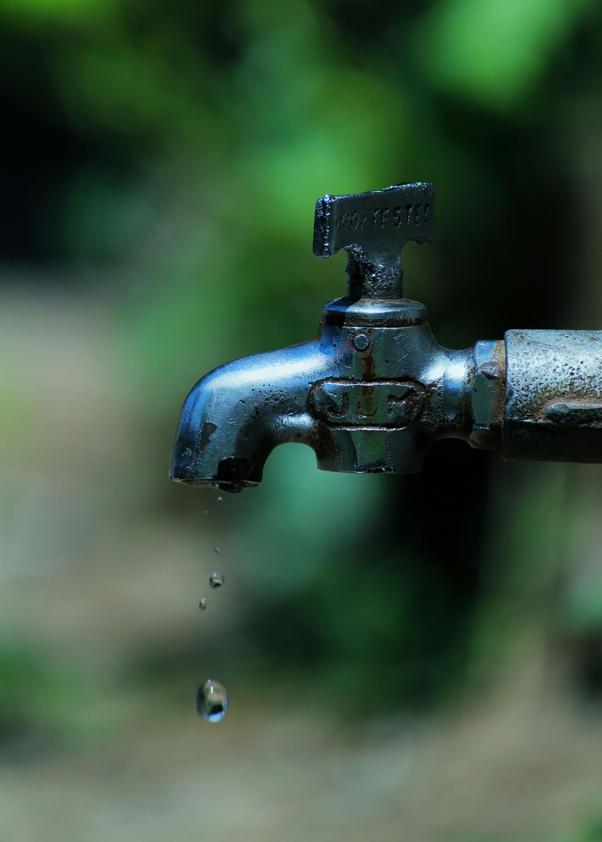 Leaking outdoor metal faucet with a water droplet against a blurred green background
