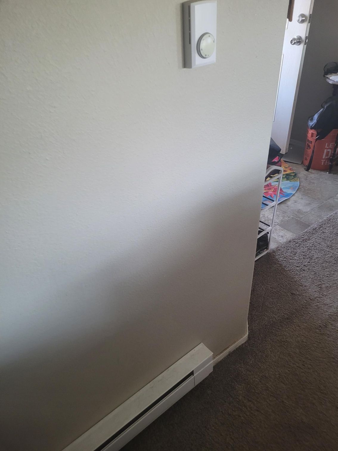 Beige wall with a white doorbell and baseboard heater, leading to a room with a door.
