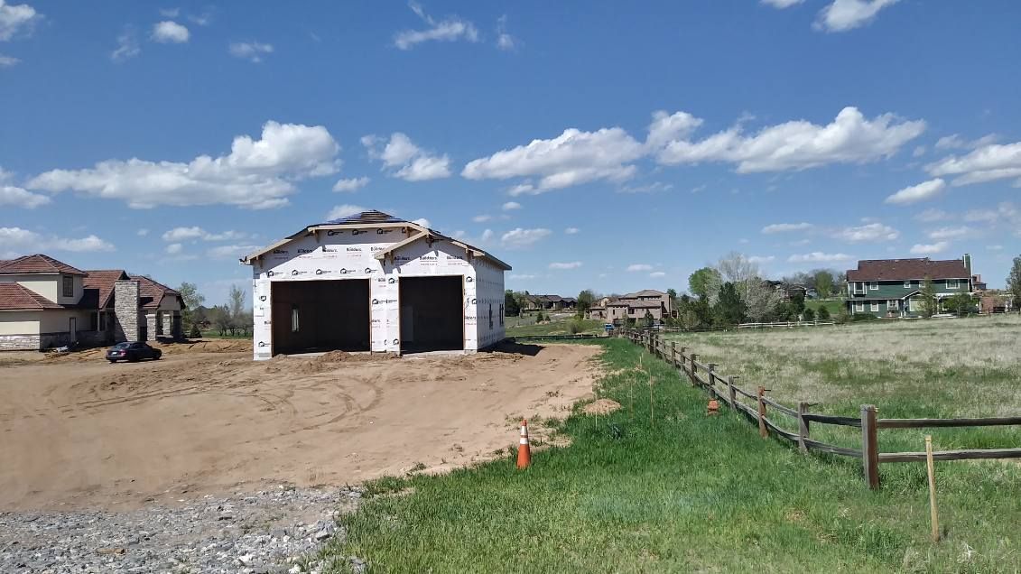 Construction site: framed garage with two bays, dirt foreground, blue sky with clouds, houses in background.