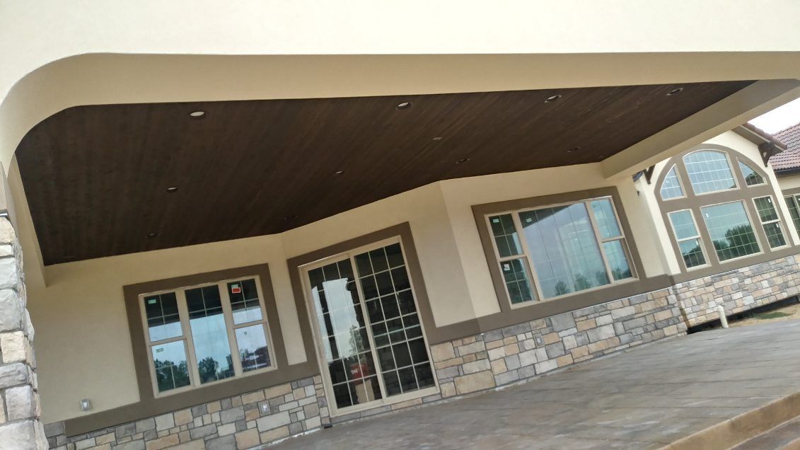 Exterior of a building with stone and beige walls, brown ceiling, large windows, and a sliding glass door.