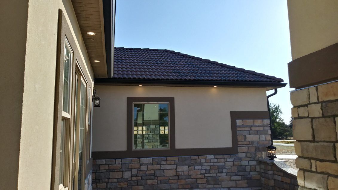 Exterior of building with dark roof, stone and tan walls, and blue sky.