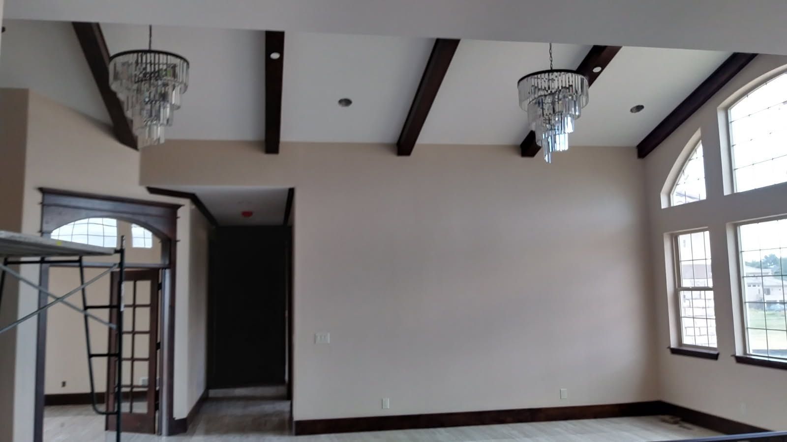 Beige room with dark wood beams, two chandeliers, and arched windows.