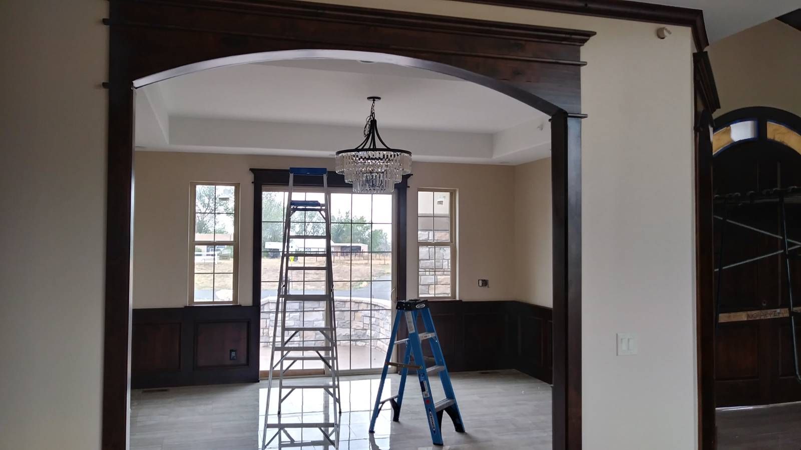 Interior room with dark wood trim and archway, windows, chandelier, ladder, and door.
