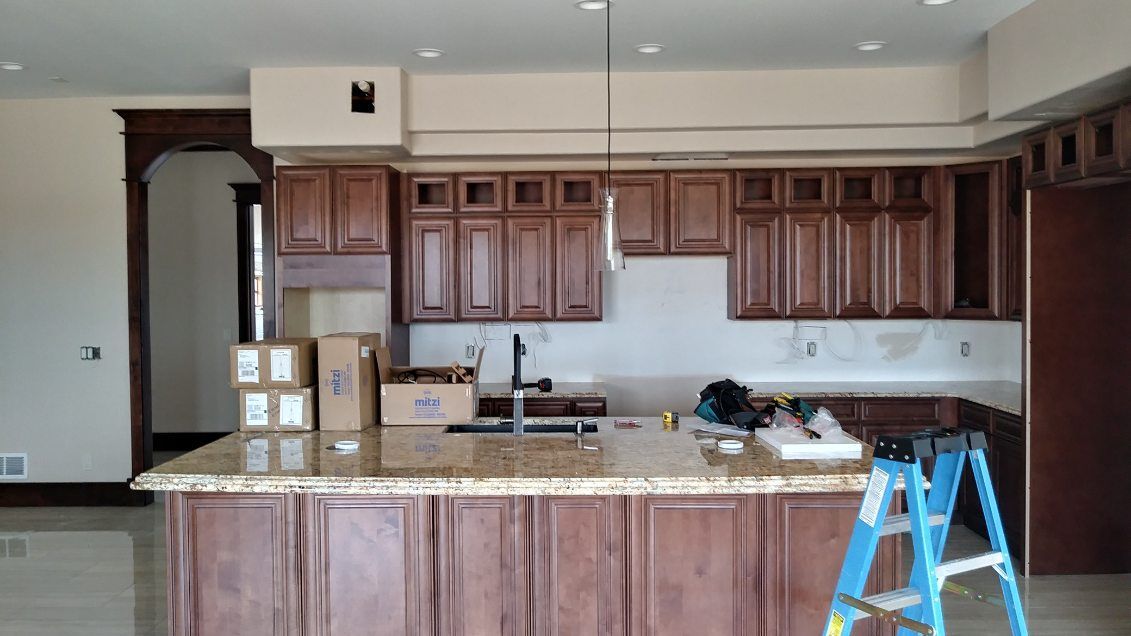 Kitchen renovation: brown cabinets, granite island, tools, and boxes; a ladder is present.