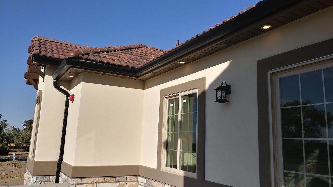 Beige stucco house with brown trim, clay tile roof, and black gutters.