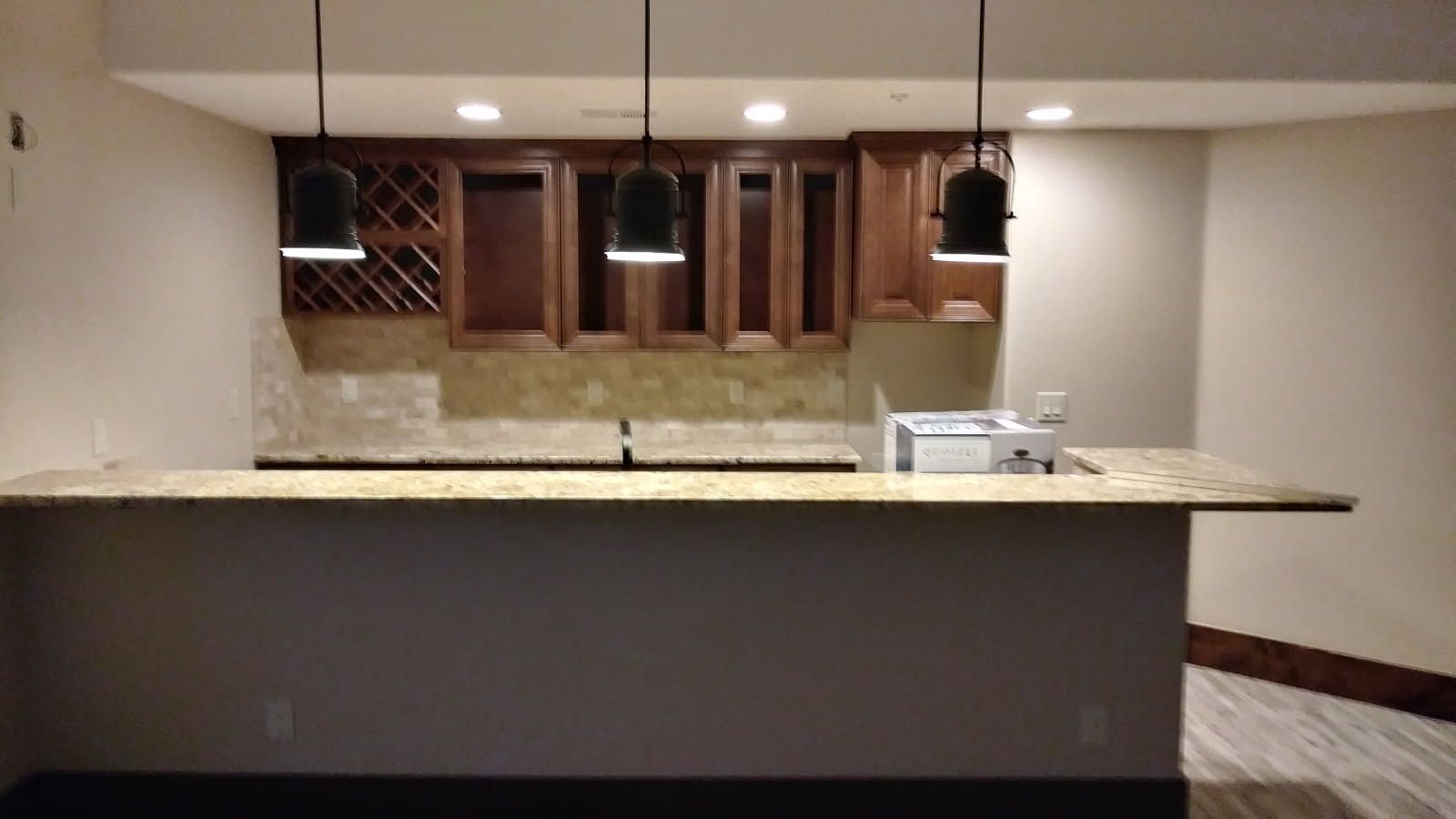 Basement bar with granite countertop, wooden cabinets, pendant lights, and tan backsplash.