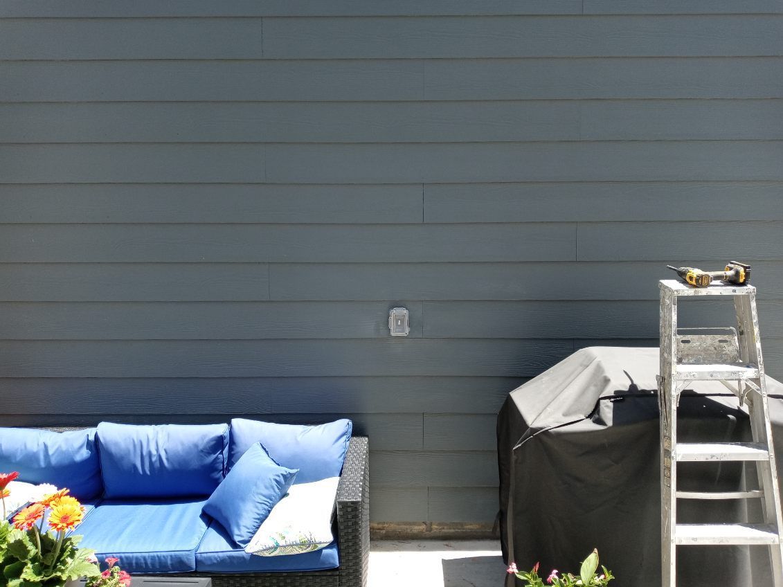 Blue outdoor sofa, gray siding, covered grill, and ladder against a building.