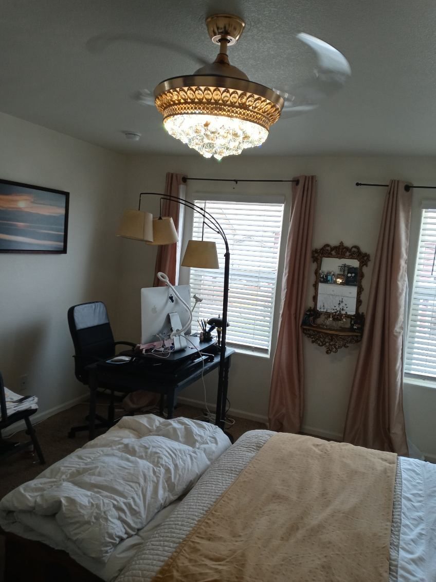 Bedroom with bed, desk, lamps, windows, gold chandelier fan, and pink curtains.