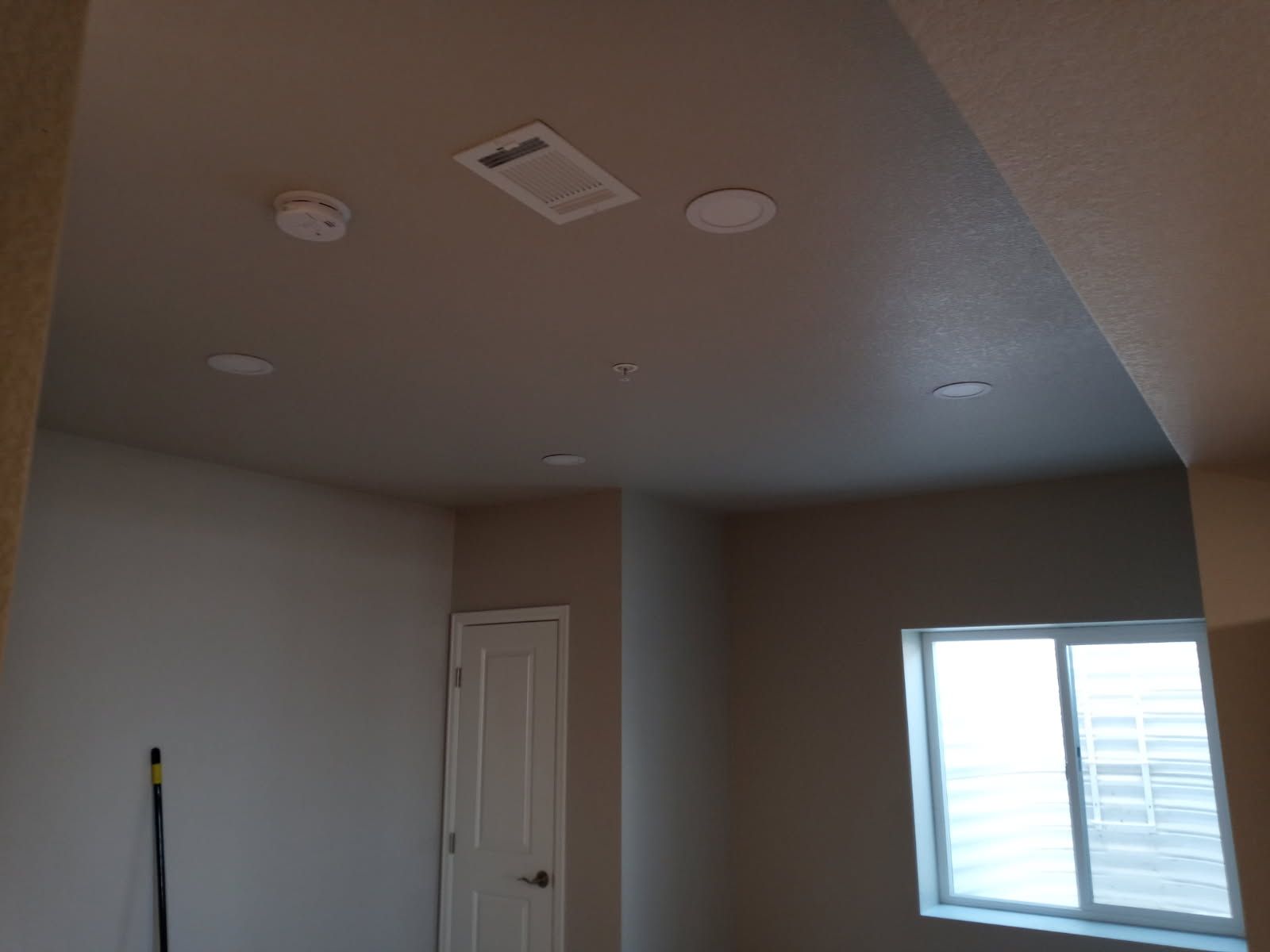 Interior view of a room with ceiling lights, smoke detector, vent, window, and door; walls are painted beige.