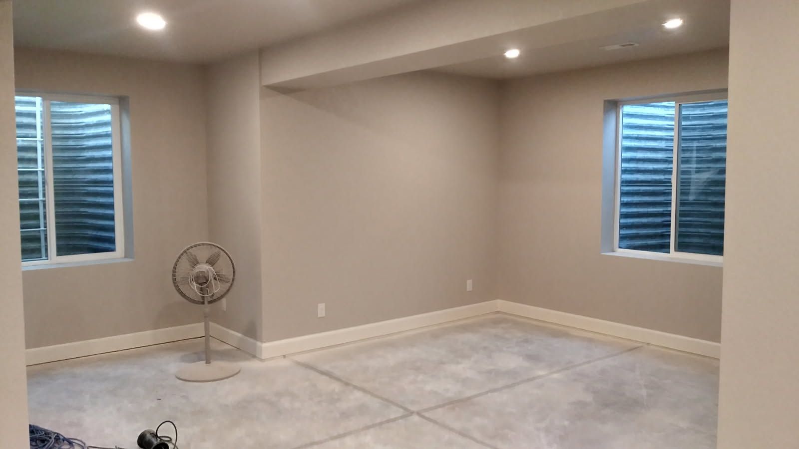 Empty room with two windows, gray walls, concrete floor, and a standing fan.