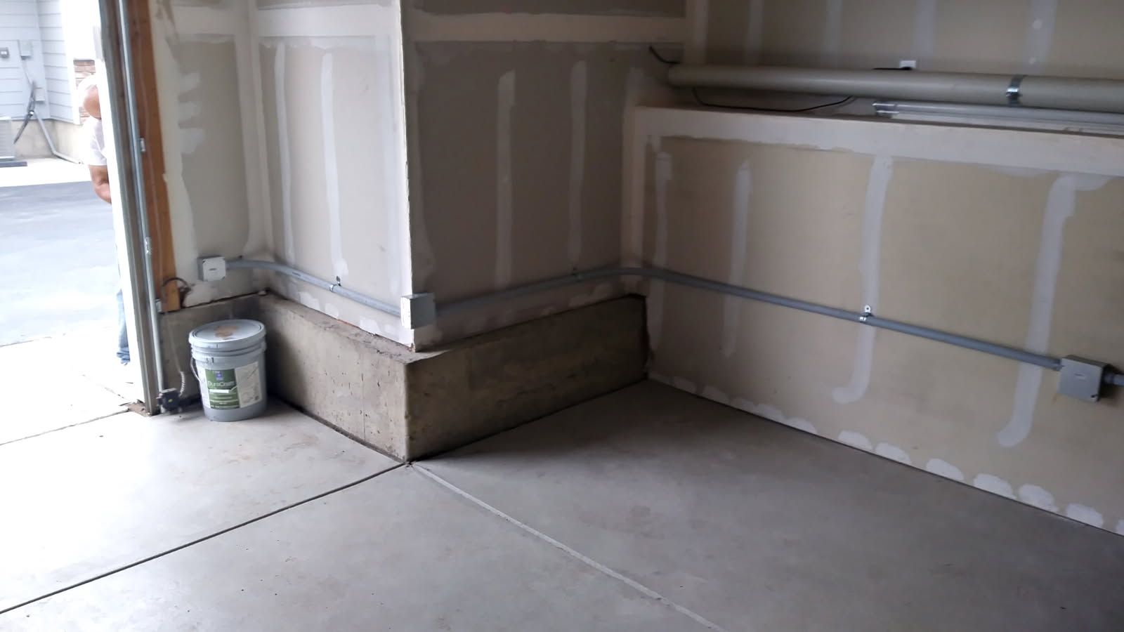Garage corner with unfinished drywall, gray floor, and a concrete base. A bucket sits near the door.