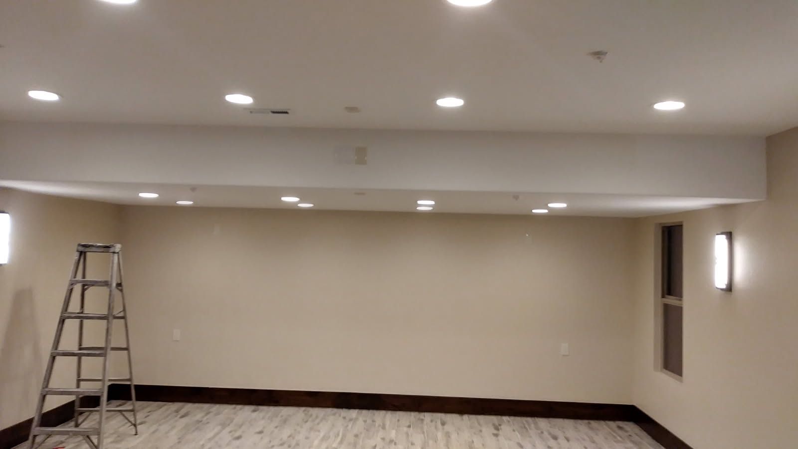 A room with recessed lighting, ladder, and two wall sconces; beige walls and wood trim.