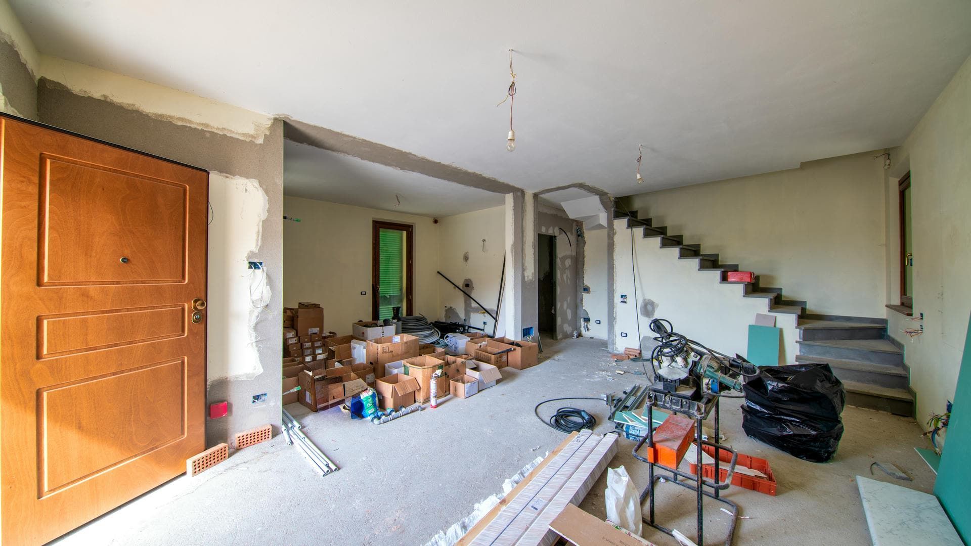 Interior during renovation, featuring a wooden door, drywall, a staircase, and construction materials.