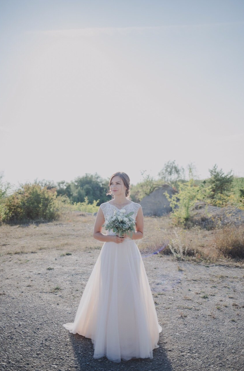 Eine Braut im Brautkleid steht mit einem Blumenstrauß in der Hand auf einem Feld.