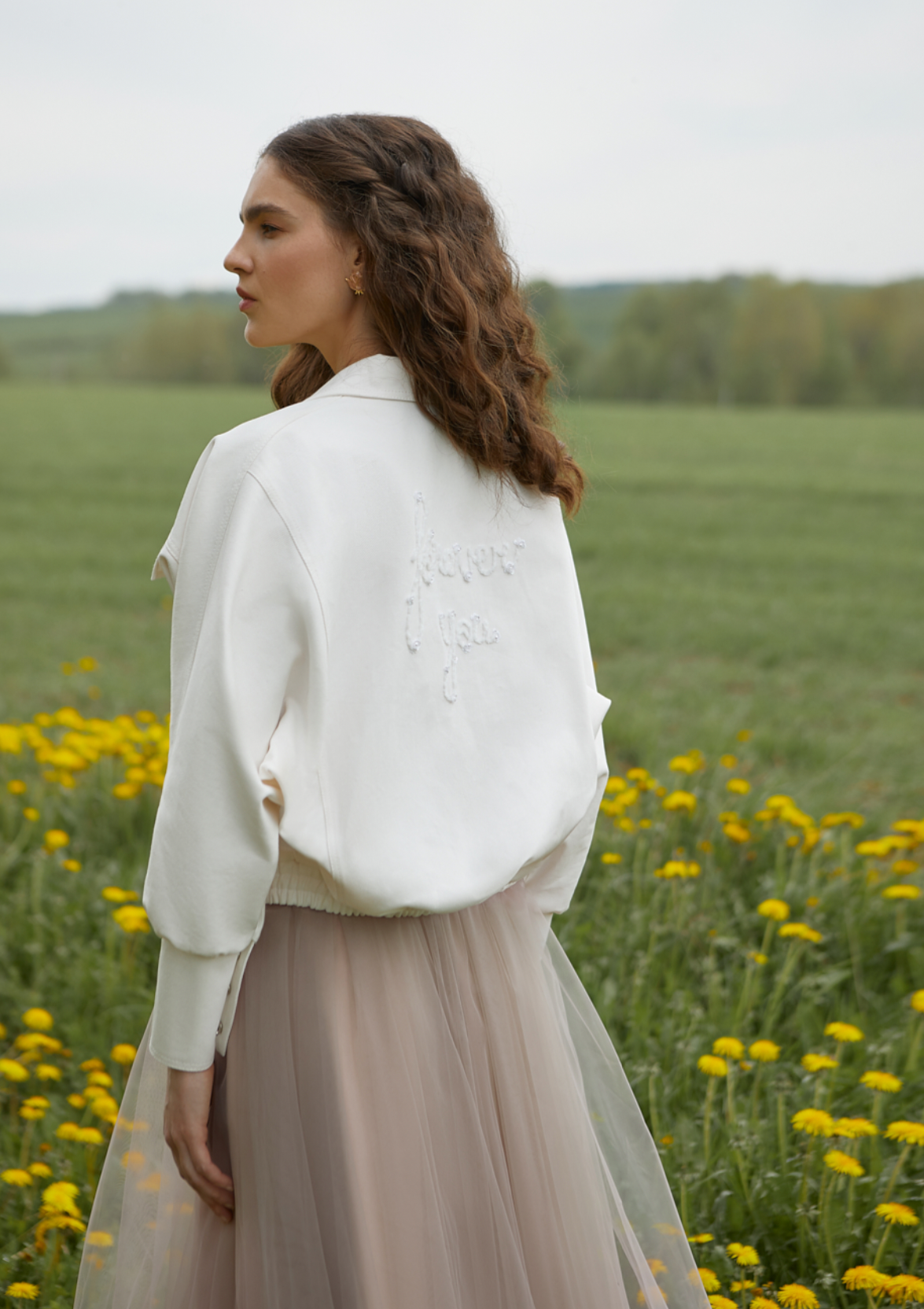 Eine Frau steht in einem Löwenzahnfeld und trägt einen weißen Pullover und einen Rock.