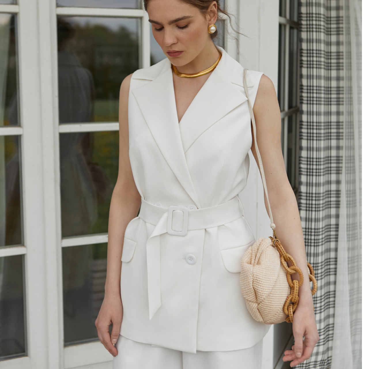 Eine Frau in weißer Weste und Hose steht vor einem Fenster.